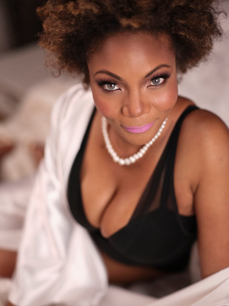 BIPOC model in black one piece lingerie with nude heels and pearls on bed in Toronto Boudoir Photography studio natural hair pink lipstick smoky eye