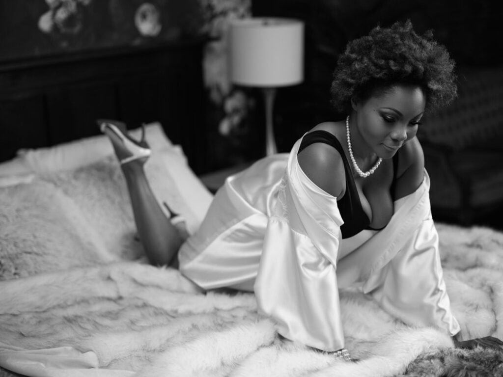Black and white image of BIPOC model with natural hair on bed wearing black lingerie pearls silk robe boudoir Toronto-Provocateur Images